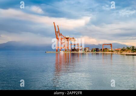 Leuchtend orangefarbene Ladekräne und gestapelte Frachtcontainer am Hafendock in Rijeka, Kroatien. Haupthafen im Osten der Stadt bei Sonnenuntergang mit schöner Aussicht Stockfoto