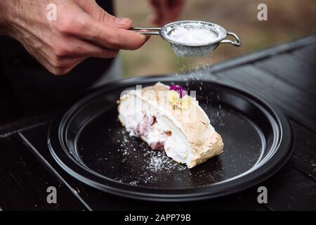 Der Koch besprüht Puderzucker auf einen leckeren und frischen Meringue roll mit Sahne, garniert mit einem Live Blume. Stockfoto