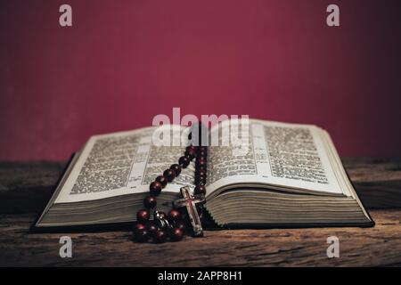 Öffnen Sie die heilige Bibel und das Kruzifix der Perlen auf einem alten Holztisch aus Eiche. Schöner roter Wandhintergrund. Stockfoto