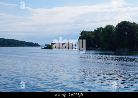 Blick auf die ruhigen Gewässer des Chiemsee (Bayern, Deutschland). In der Ferne befinden sich ein Holzhäuschen und eine bewaldete Insel. Der Himmel ist sehr blau. Stockfoto