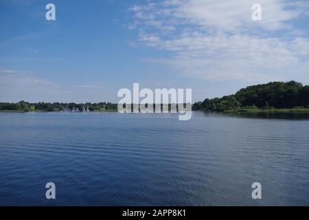 Blick auf die ruhigen Gewässer des Chiemsee (Bayern, Deutschland). In der Ferne befinden sich ein Hafen und bewaldete Inseln. Der Himmel ist sehr blau. Stockfoto