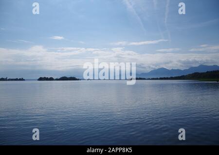 Blick auf die ruhigen Gewässer des Chiemsee (Bayern, Deutschland). In der Ferne befinden sich Berge und bewaldete Inseln. Der Himmel ist sehr blau. Stockfoto