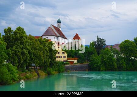 Panorama auf einen undurchsichtigen blauen Fluss, der durch eine kleine Stadt führt, mit mehreren Gebäuden und einer Kirche. Das Flussufer ist von grünen lebhaften Bäumen überwuchert. Stockfoto