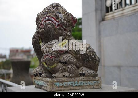 Chinesischer Schutzlöwe vor dem Grab auf einem Friedhof. Bunte Steinskulptur, die seine Zähne zeigt, mit einer Pfote auf einem Ball. Religiös und mystisch. Stockfoto