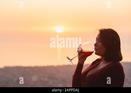 Frau mit Rotwein in den Bergen des Sonnenuntergangs, Nahaufnahme von Hand mit Glas Wein. Elegante Frau, die bei Sonnenuntergang die schöne Berglandschaft der Berge und das Meer genießt Stockfoto