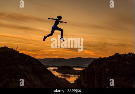Frau springt durch die Kluft zwischen Hill.Woman, die über eine Klippe auf dem Hintergrund des Sonnenuntergangs springt, Idee des Geschäftskonzepts Stockfoto