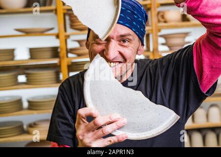 Lustiger Töpfer lächelt durch die zerbrochenen Keramikplatten in seinem Atelier ih vor Töpferkunstprodukten, Kleinkunstunternehmerkonzept Stockfoto