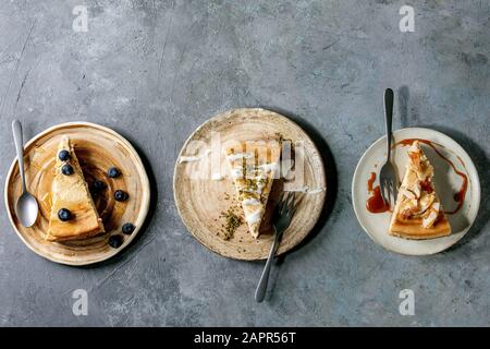 Käsekuchen mit verschiedenen Belägen und Dekor auf Keramikplatten mit Gabeln in Reihe über grauem Texturhintergrund. Flaches Layout, Platz Stockfoto