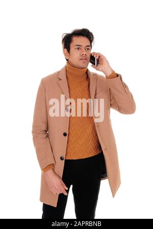 Ein junger Mann in einem beigefarbenen Wintermantel, der auf seinem Handy steht und spricht und einen Pullover trägt, isoliert für weißen Hintergrund Stockfoto
