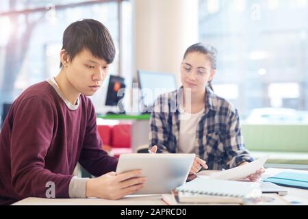 Asiatischer junger Mann, der zusammen mit einer jungen Frau am Tisch sitzt und digitale Tabletts studiert und verwendet Stockfoto