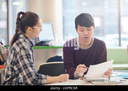 Asiatischer junger Mann liest einen Bericht und diskutiert ihn zusammen mit einer jungen Frau, während er im Klassenzimmer am Tisch sitzt Stockfoto