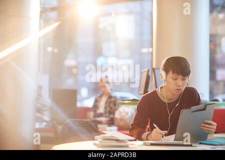 Asiatischer junger Mann in Kopfhörern, der mit Zwischenablage am Tisch sitzt und Notizen in Notizbuch macht, das er in der Bibliothek arbeitet Stockfoto