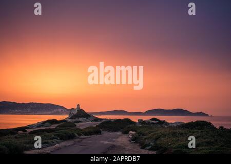 Oranger und violetter Himmel bei Sonnenuntergang über den alten Ruinen eines genuesischen Turms in Punta Caldanu bei Lumio und der Zitadelle von Calvi in der Balagne o Stockfoto