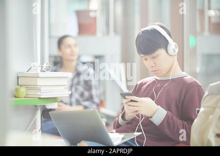 Student der asiatischen Hochschule in Kopfhörern sitzt auf Sofa mit Laptop auf den Knien und mit Handy Stockfoto