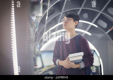 Asiatische Studenten in Brillen, die Bücher halten und Bücherregal betrachten, während sie in der modernen Bibliothek stehen Stockfoto