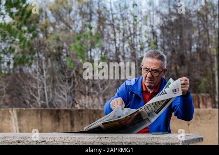 Alter Mann mit Brille liest die Zeitung im Park mit den Bäumen im Hintergrund Stockfoto