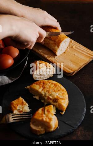 Spanisches Kartoffelomelett in Scheiben geschnitten und mit Brot serviert. Eine Frau schneidet im Hintergrund einige Brotscheiben. Stockfoto