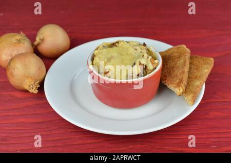 Zwiebelsuppe aus Gratin mit Brot und Käse auf rotem Holztisch mit Zwiebeln daneben als traditionelles herbstliches Essen Stockfoto