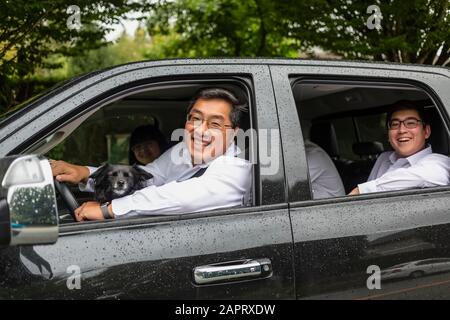 Die Familie fährt im Auto mit dem Hund, dem fahrenden Vater und den erwachsenen Kindern auf dem Rücksitz, schaut auf die Kamera und lächelt Stockfoto