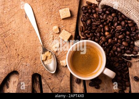 Auf dem rustikalen Holztisch, im Vordergrund, eine Tasse Espresso-Kaffee, brauner Zucker und gebratene Kaffeebohnen. Stockfoto