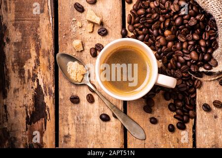 Auf dem rustikalen Holztisch, im Vordergrund, eine Tasse Espresso-Kaffee, brauner Zucker und gebratene Kaffeebohnen. Stockfoto