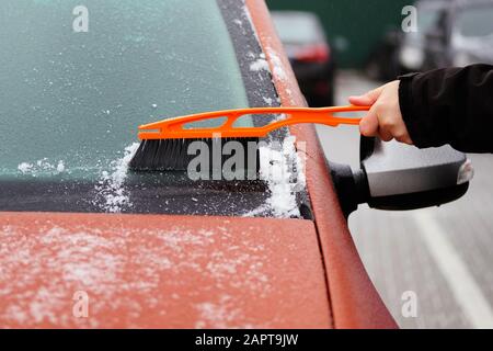 Der Mann räumt Schnee aus eisigen Autofenstern. Orangefarbene Bürste in bemannt Hand. Windschutzscheibe des Wagens, horizontale Ansicht. Stockfoto