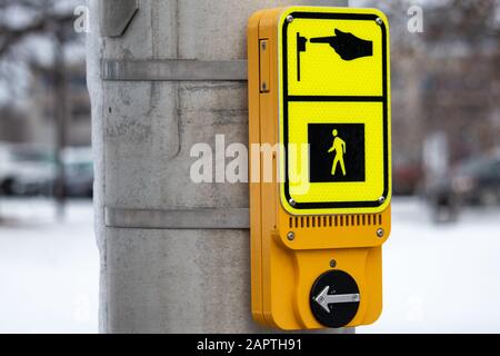 Eine moderne gelbe Fußgängerruftaste ist auf einem Metall-Verkehrssignalpol montiert und im Winter zu sehen. Die Taste trägt ein Schild, das ihren Zweck angibt Stockfoto