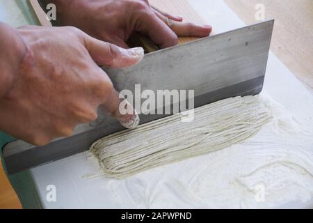 Die Hand eines Mannes hält ein großes Metall-Nudelmesser, um Soba-Nudeln auf einem Schneidebrett auf einer Schicht Buchweizenmehl zu schneiden. Stockfoto