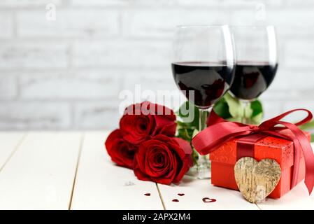 Valentinstag Konzept. Zwei Gläser Wein, rote Rosen, Geschenkbox, Holzherz und brennende Kerzen auf einem weißen Holztisch. Selektiver Fokus. Stockfoto