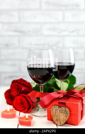 Valentinstag Konzept. Zwei Gläser Wein, rote Rosen, Geschenkbox, Holzherz und brennende Kerzen auf einem weißen Holztisch. Selektiver Fokus. Stockfoto