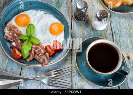 Das Frühstück besteht aus Spiegelei, Speck, roher Tomate, Toast auf der alten blauen Porzellanplatte und einer Tasse schwarzen Kaffee auf dem alten Holztisch Stockfoto