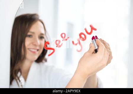 Hand der fröhlichen jungen Brunette-Frau, die mit Lippenstift auf Spiegel schreibt Stockfoto