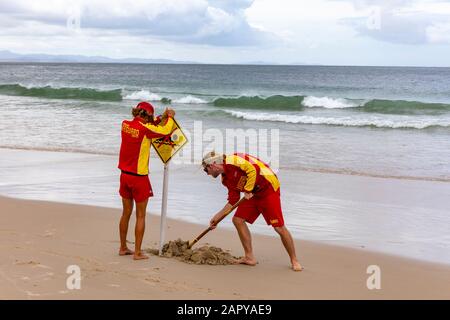 Zwei Rettungsschwimmer graben ein Loch am Strand, um während eines stürmischen Sommers, Australien, in Byron Bay das Schild ohne Schwimmzeichen zu Pflanzen Stockfoto