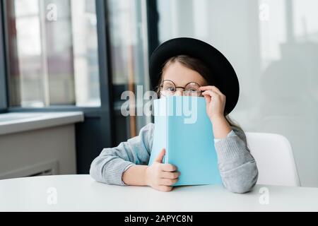 Süßes kleines Schulmädchen in Brillen und Hut aus offenem Buch Stockfoto