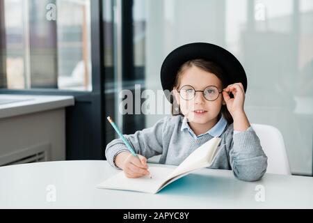 Liebenswürdige, clevere Schulmädchen im Hut, die Sie beim Erstellen von Notizen in einem Werbebuch betrachtet Stockfoto