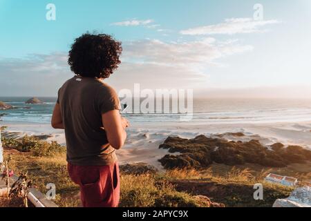 Mann mit lockigen Haaren, der das Meer, umgeben von Felsen, beobachtet Und Grün unter dem Sonnenlicht Stockfoto