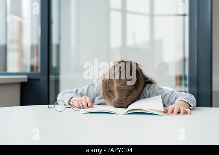 Sehr müde oder gelangweilte Schulmädchen mit Brillen, die auf einem offenen Werbebuch am Schreibtisch einschnallen Stockfoto