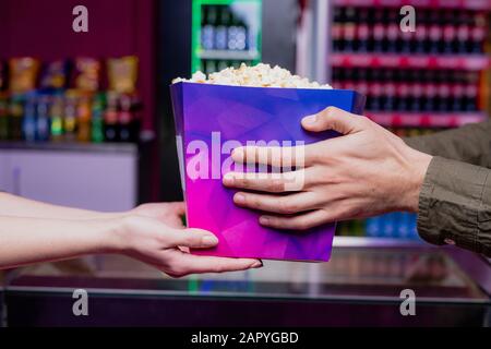 Hände des jungen Mannes, der eine große lila Papierbox voller Popcorn im Kino nimmt Stockfoto