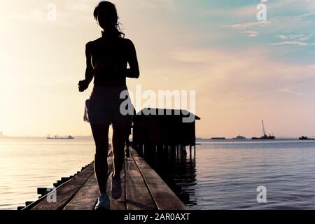 Silhouette der Frau, die morgens auf einer Holzbrücke bei Sonnenaufgang joggt. Stockfoto