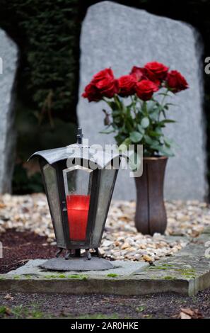 Grablaterne mit brennender Kerze auf einem Grab vor einem Blumenstrauß roter Rosen in verschwommenem Hintergrund Stockfoto