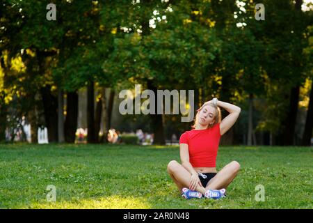 Junge schöne Frau, die im Park auf dem Gras sitzt und Muskeln trainiert und ausdehnt. Stockfoto