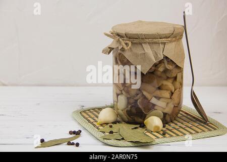 Vorgegärte Champignons in einem Glas Knoblauchzopf mit schwarzem Pfefferlöffel auf hellem Holzhintergrund Stockfoto