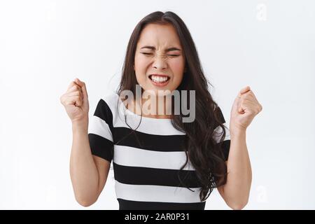 Gestresste und ängstliche junge panickende asiatische Brunette-Frau im gestreiften T-Shirt, die geklemmte Fäuste aufhebt und sie nervös schüttelt, als enge Augen und Knirschen Stockfoto