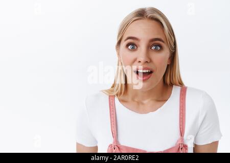 Nahaufnahme schockiert und beeindruckte blonde Mädchen mit blauen Augen in rosafarbenen Overalls, T-Shirt, offenem Mund und Stare fasziniert fanden erstaunliche Nachrichten heraus, stehend Stockfoto