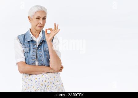 Durchsetzungsstarke, selbstbewusste, stilvolle Seniorin, Großmutter in trendiger Weste, Kleid, mit einem Zeichen in Ordnung, Augenzwinkern, die alles unter Kontrolle haben, bewerten Sie etwas Stockfoto