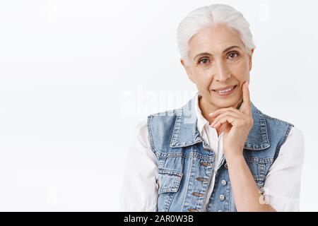 Close-up Portrait niedlich, modern charmante Seniorin mit gekämmtem grauem Haar in Denim-Weste, leicht anrührend und lächelnd, mit Antifaltenbeschlag Stockfoto