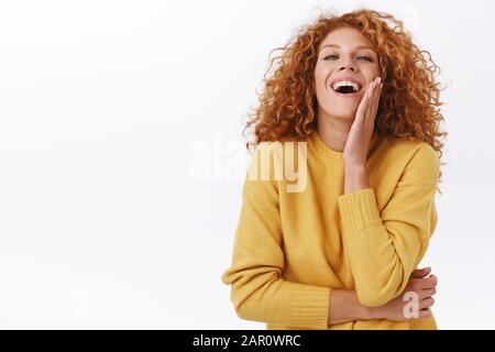 Attraktive sassy und feminine Rotkopffrau mit lockigen Haaren, tragen gelben Pullover, lachen arrogant und angetan, rührende Wange heben Kopf auf und Stockfoto