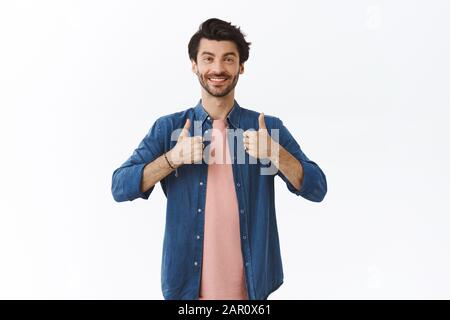 Fröhlicher, aufrüttiger, gut aussehender bärtiger kaukasischer Mann im Hemd, T-Shirt mit Daumen nach oben und lächelnd, geben Zustimmung, unterstützen Sie bei der Herstellung exzellenter Stockfoto