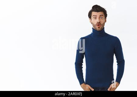 Überraschte einige bärtige männliche Unternehmer im blauen Pullover mit hohem Hals, lächelnd gewundert, beeindruckt und staunend, sehen Sie sich etwas tolles an Stockfoto