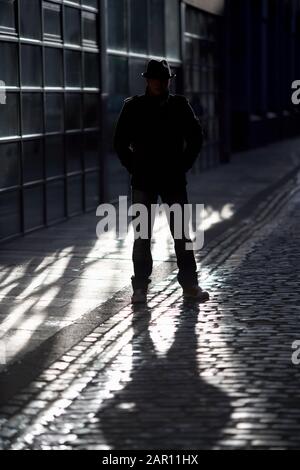 Geheimnisvoller Mann mit Hut und Mantel, der im Sonnenlicht auf einer alten gepflasterten Straße silhouettiert steht Stockfoto